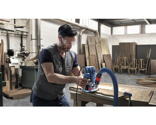 Muž s ochrannými brýlemi opracovává dřevo horní frézkou v dílně.