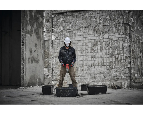 Osoba s ochrannou přilbou a obličejovou maskou míchá materiál míchadlem ve stavebním prostředí.