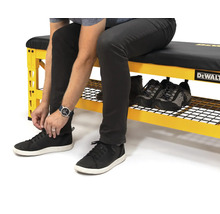 Osoba sedí na žluté pracovní lavici s úložným prostorem na boty a zavazuje si boty. Na pracovní lavici jsou další boty. Logo DeWalt