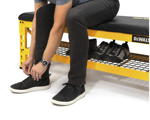 Osoba sedí na žluté pracovní lavici s úložným prostorem na boty a zavazuje si boty. Na pracovní lavici jsou další boty. Logo DeWalt