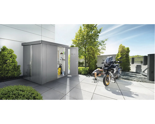 Zahradní scéna s kůlnou na nářadí, ve které je vidět tlaková myčka a police. Před kůlnou na nářadí opravuje muž motorku.