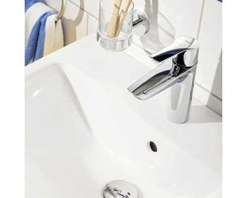 Fotografie bílého umyvadla s lesklou vodovodní baterií, kelímkem na zubní kartáčky a držákem na ručníky