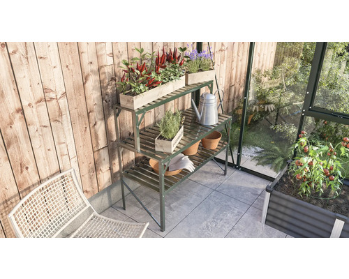 Zahradní scéna s kovovým stolem na rostliny, osázenými dřevěnými bednami, konví a zahradním nářadím ve skleníku