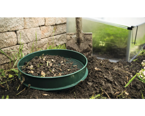 Zahradní síto s hlínou, lopata a skleník v pozadí