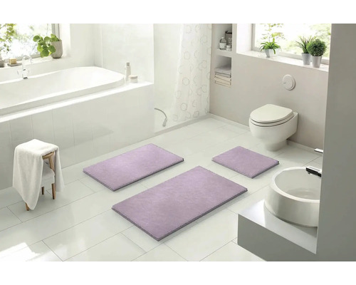 Světlá koupelna s vanou, toaletou, umyvadlem a sadou tří fialových koupelnových předložek.