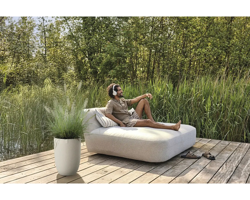 Muž relaxuje na zahradním lehátku se sluchátky, vedle něj květináč s okrasnou trávou na dřevěné terase.