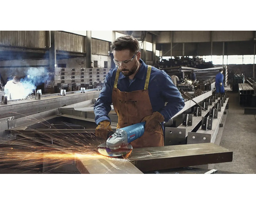 Muž s ochrannými brýlemi a koženou zástěrou obsluhuje úhlovou brusku v dílně.