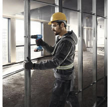 Stavební dělník s ochrannou helmou a brýlemi používá akumulátorový šroubovák na kovové konstrukci.