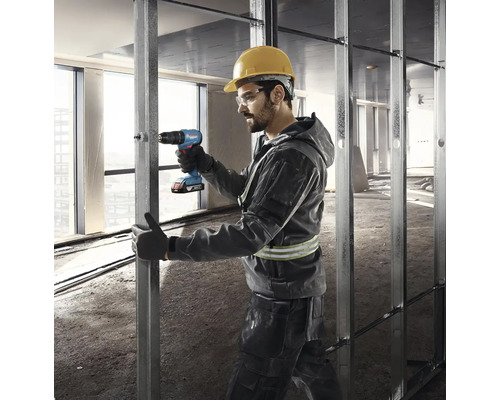 Stavební dělník s ochrannou helmou a brýlemi používá akumulátorový šroubovák na kovové konstrukci.