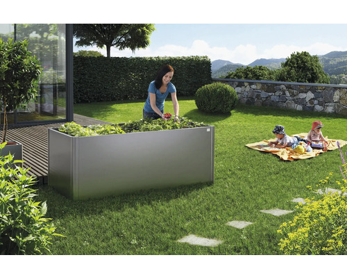 Zahradní scéna s vyvýšeným záhonem, žena sklízí zeleninu, děti si hrají na dece na zahradě.