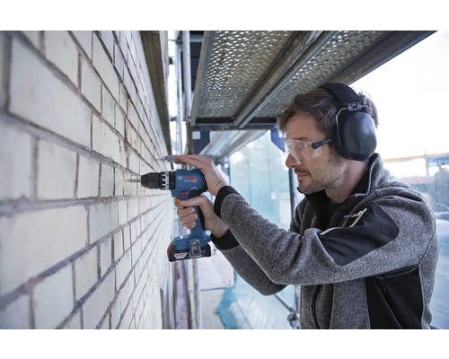 Muž vrtá aku vrtačkou Bosch do cihlové zdi, má ochranné brýle a chrániče sluchu.