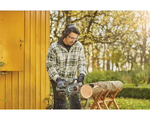 Muž řeže kus dřeva na kozlíku pomocí akumulátorové řetězové pily, přičemž má ochranné brýle a sluchátka.