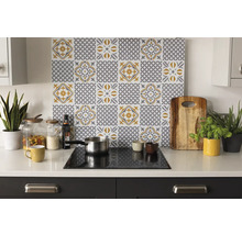 Organizovaná světlá kuchyně se zadní stěnou s šedo-žlutým vzorem dlaždic nad varnou deskou na bílé pracovní desce s doplňky a rostlinami.