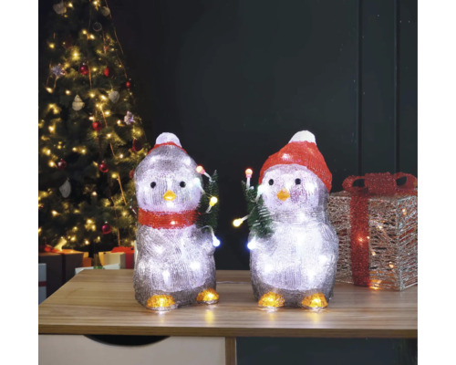 Dvě figurky tučňáků s osvětlením a vánočním motivem na stole před vánočním stromkem.