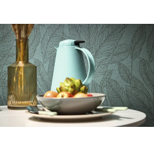 Dekorativní zátiší s vázou, termoskou a mísou ovoce před tapetou se vzorem listů.