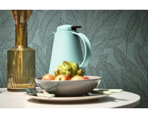 Dekorativní zátiší s vázou, termoskou a mísou ovoce před tapetou se vzorem listů.