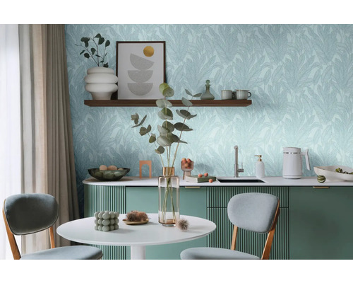 Světlá místnost se stolem, židlemi a kuchyňským koutem před stěnou s tapetou s listovým motivem.