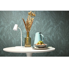 Dekorativní jídelní stůl s lampou, termoskou, vázou se suchými květinami a mísou s ovocem před tapetou se vzorem listů