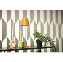 Dekorativní scéna s lampou, svíčkami a hrnkovou rostlinou na polici před geometrickou tapetou.