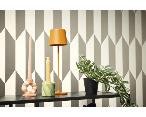 Dekorativní scéna s lampou, svíčkami a hrnkovou rostlinou na polici před geometrickou tapetou.