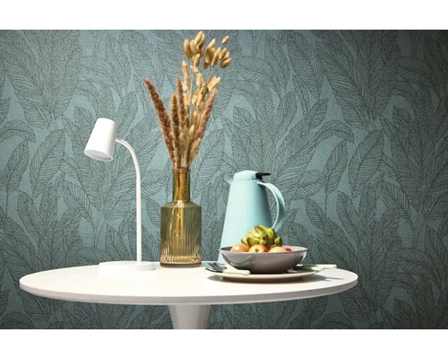 Stůl s lampou, vázou, termoskou a mísou před tapetou s listovým vzorem.