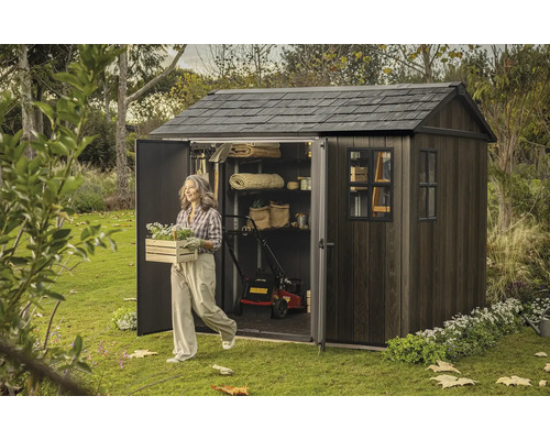 Zahradní scéna s kůlnou na nářadí, sekačkou na trávu a ženou nesoucí truhlík s květinami.