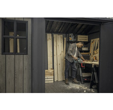 Muž řeže dřevo v kůlně s nářadím a uskladněným dřevem.