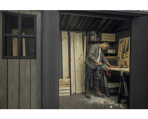 Muž řeže dřevo v kůlně s nářadím a uskladněným dřevem.