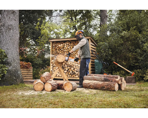 Muž řeže dřevo motorovou pilou před kůlnou na dřevo na zahradě.