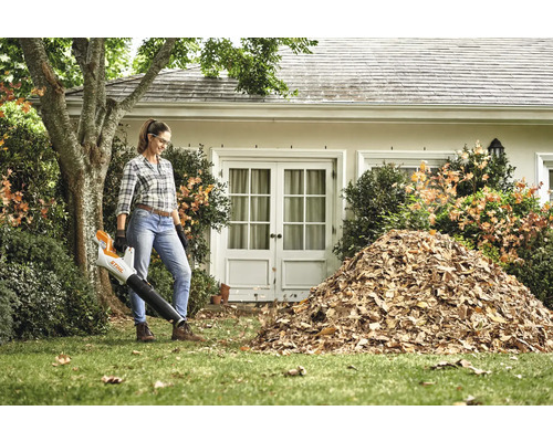 Žena používá foukač listí na zahradě před domem s hromadou listí