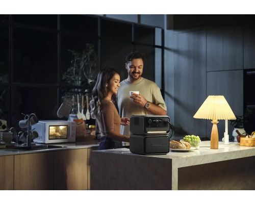 Pár v kuchyni s přenosným generátorem energie, který napájí lampu, zatímco v pozadí běží mikrovlnná trouba.