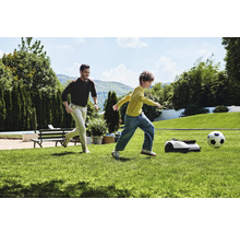 Muž a dítě hrají fotbal s robotickou sekačkou na zahradě.