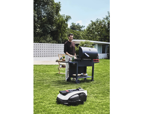 Muž griluje na zahradě s grilem a robotickou sekačkou.
