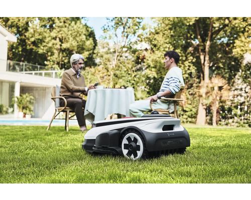 Scéna s robotickou sekačkou na trávu na zahradě, zatímco dvě osoby sedí u stolu