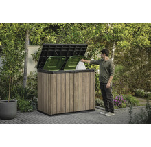 Dřevěný box na popelnice se dvěma zelenými popelnicemi a osobou, která do něj vhazuje pytel s odpadky