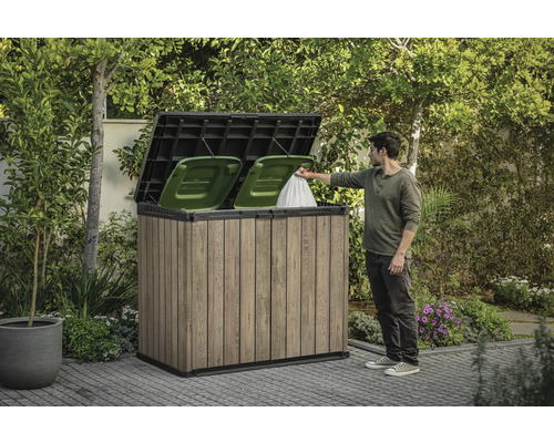 Dřevěný box na popelnice se dvěma zelenými popelnicemi a osobou, která do něj vhazuje pytel s odpadky