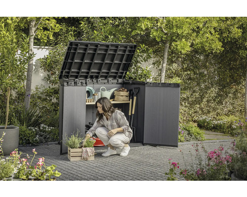 Zahrada se ženou klečící před otevřenou zahradní skříní, ve které je uloženo zahradní nářadí.