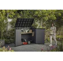 Zahradní scéna se ženou nesoucí truhlík s rostlinami vedle kůlny na nářadí plné zahradního nářadí a sekačky na trávu