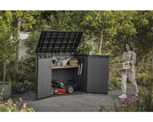 Zahradní scéna se ženou nesoucí truhlík s rostlinami vedle kůlny na nářadí plné zahradního nářadí a sekačky na trávu