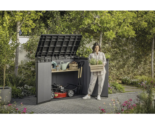Zahradní scéna s domkem na nářadí, zahradním nářadím a ženou držící bednu s rostlinami.