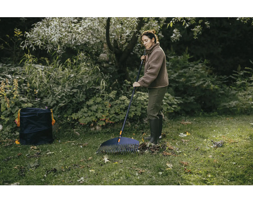 Žena shrabuje listí na zahradě hráběmi na listí a zahradním pytlem.