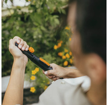 Osoba drží zahradní hadici s rozprašovací hlavicí na pozadí zelených rostlin a žlutých květů.