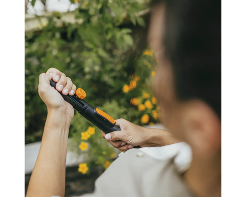 Osoba drží zahradní hadici s rozprašovací hlavicí na pozadí zelených rostlin a žlutých květů.