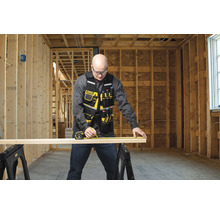 Řemeslník měří dřevěnou desku pomocí svinovacího metru na dvou kozách v hrubé stavbě.