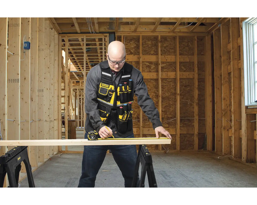 Řemeslník měří dřevěnou desku pomocí svinovacího metru na dvou kozách v hrubé stavbě.