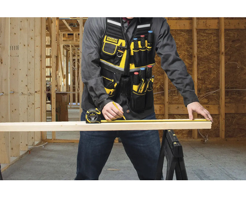 Řemeslník měří dřevěnou desku pomocí svinovacího metru a tužkou, má na sobě pracovní vestu