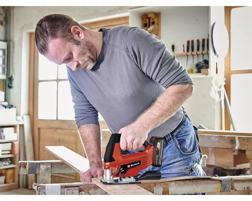 Muž řeže dřevěnou desku akumulátorovou přímočarou pilou Einhell v dílně.