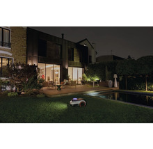 Robotická sekačka na trávu při noční práci v zahradě s domem a bazénem.