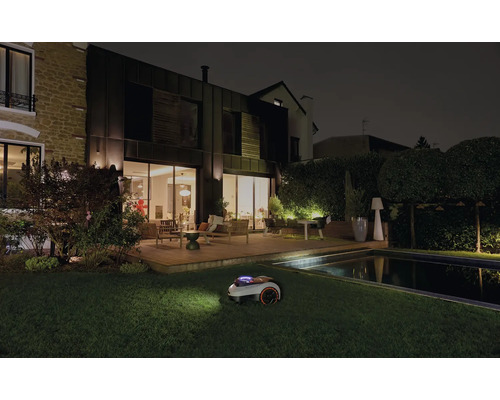 Robotická sekačka na trávu při noční práci v zahradě s domem a bazénem.