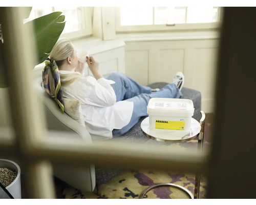 Žena relaxuje s nápojem na pohovce, v popředí kbelík s produktem Abamal na stole